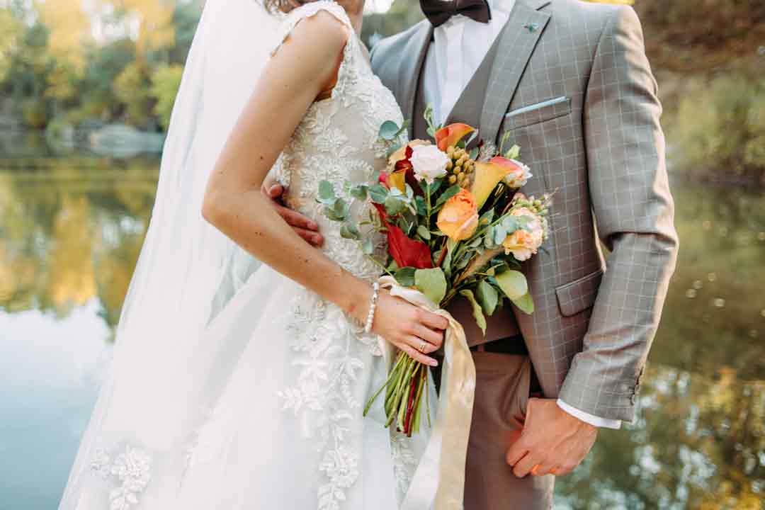 Close up of bride and groom, bride holds autumnal wedding flowers and wears a lace dress, groom wears a grey checked suit